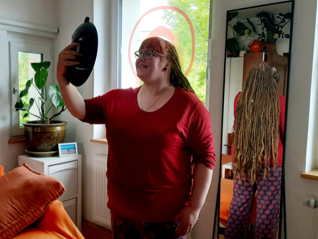 Rike mit dem Handspiegel vor dem großen Spiegel, um ihre Dreadlocks von hinten zu betrachten.