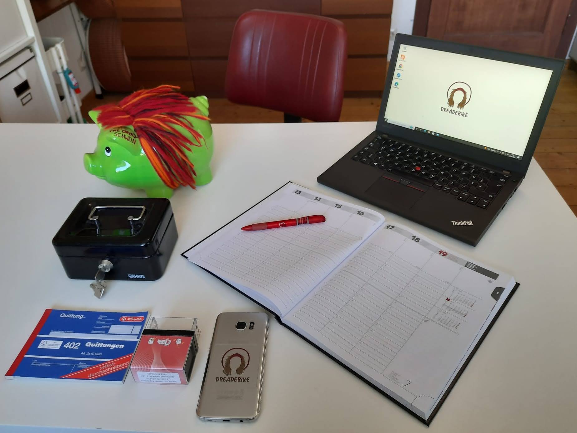 DREADERikes Business-Bereich: Ein Schreibtisch, auf dem ein Laptop mit dem DREDERike Logo steht. Außerdem liegen ein Kalender, ein roter Kugelschreiber, ein Handy mit dem DREADERike Logo, eine Kasse, Quittungsblock, Stempel und ein Sparschwein mit Wolldreadlocks auf dem Tisch. Vor dem Tisch steht ein dunkelroter Retro-Friseurstuhl.